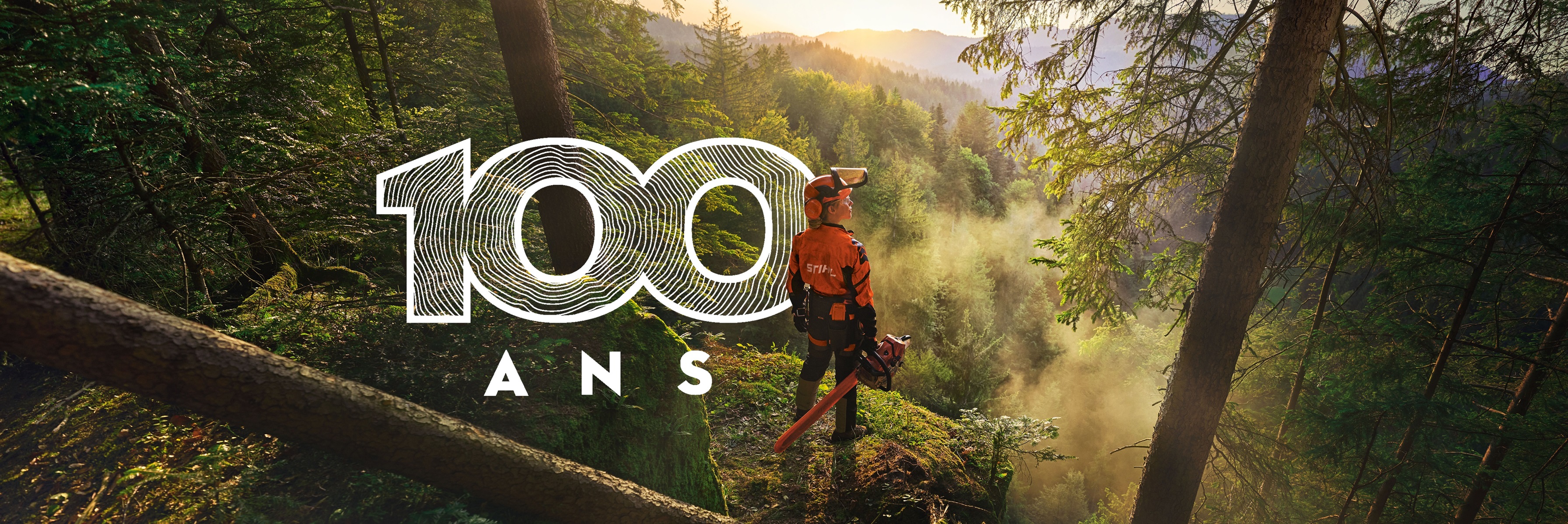 Une femme se tient au bord d'une falaise dans une forêt d'un vert saturé. Elle porte des vêtements de travail STIHL orange, le casque ADVANCE ProCom et tient une tronçonneuse STIHL MS.