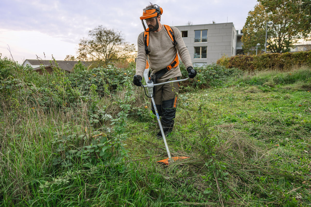 Ein Mann in STIHL Schutzausrüstung mäht mit einem Akku-Freischneider STIHL FSA 200 eine wild überwucherte Grünfläche