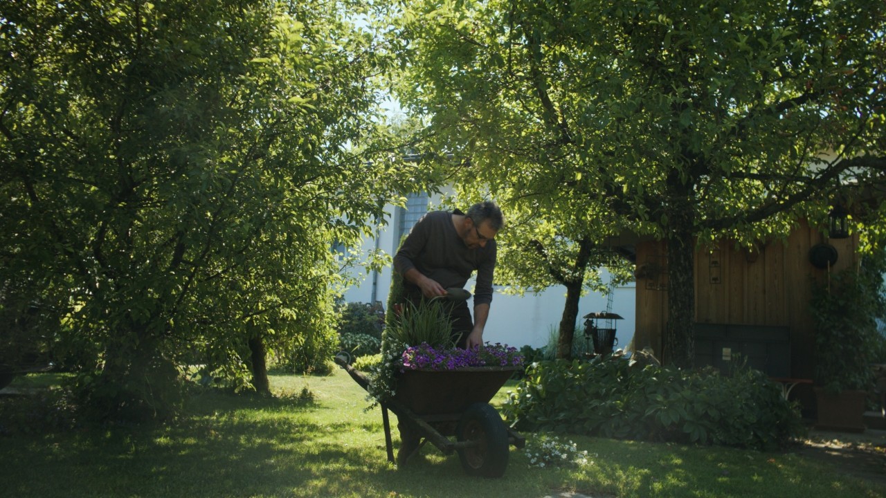Rolf arbeitet im Garten mit einer Schaufel an Blumen in einer Schubkarre, grüne Rasenfläche und Pflanzen im Hintergrund, Real People. STIHL People. Kampagne