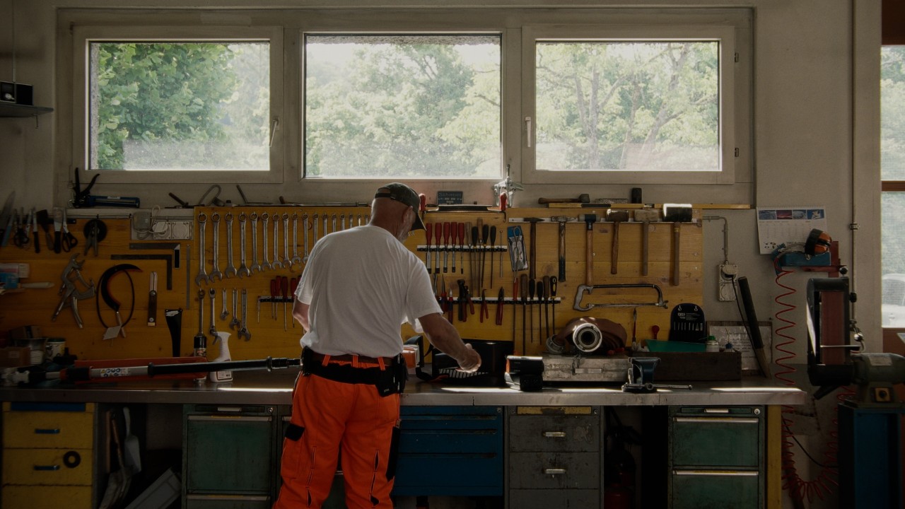 Lukas in einer Werkstatt von hinten zu sehen, hält einen STIHL Akku und nimmt ihn aus einem STIHL Akku‑Heckenschneider auf der Werkbank heraus, Real People. STIHL People. Kampagne