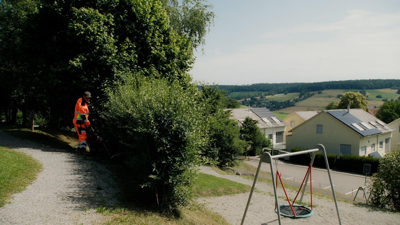 Lukas bei der Pflege einer Hecke mit STIHL Heckenschneider in einer öffentlichen Grünanlage, im Hintergrund ein Spielplatz, Real People. STIHL People. Kampagne