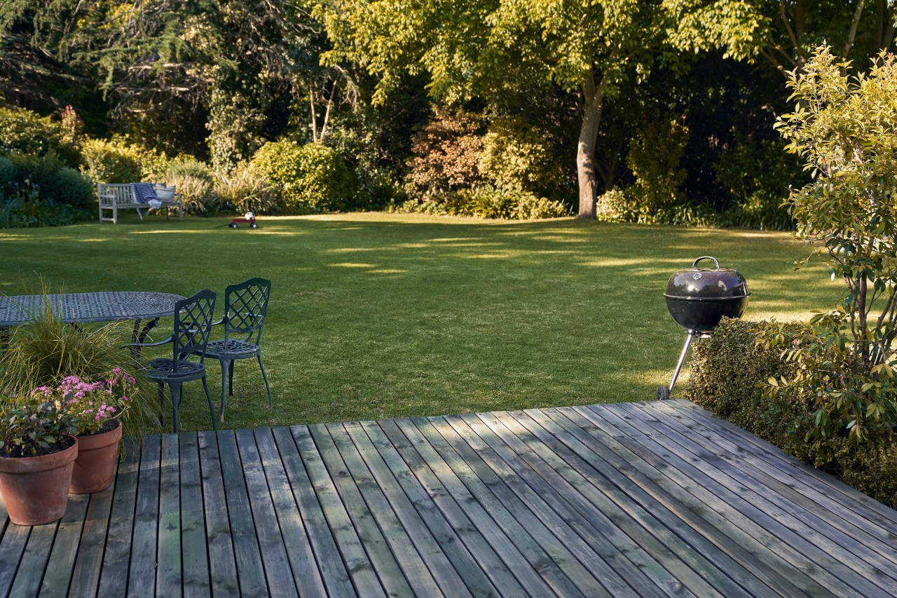 Nettoyage de terrasse en bois devant un jardin verdoyant avec barbecue, chaises et pots de fleurs