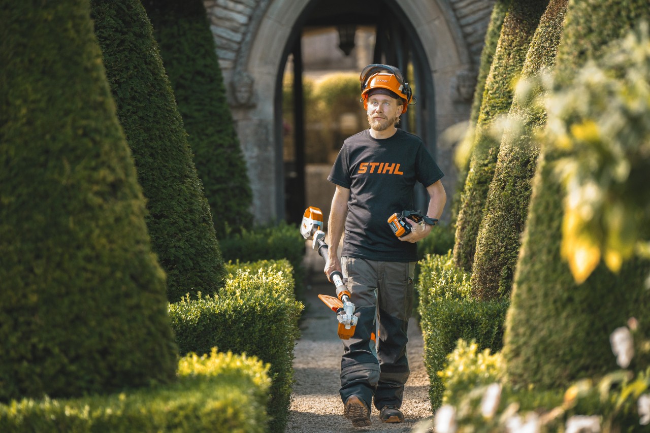 Le chef jardinier Andrew Wain traverse une allée du jardin, équipé d'un casque de protection, d'un t-shirt STIHL et d'un outil à batterie STIHL avec une batterie séparée à la main.