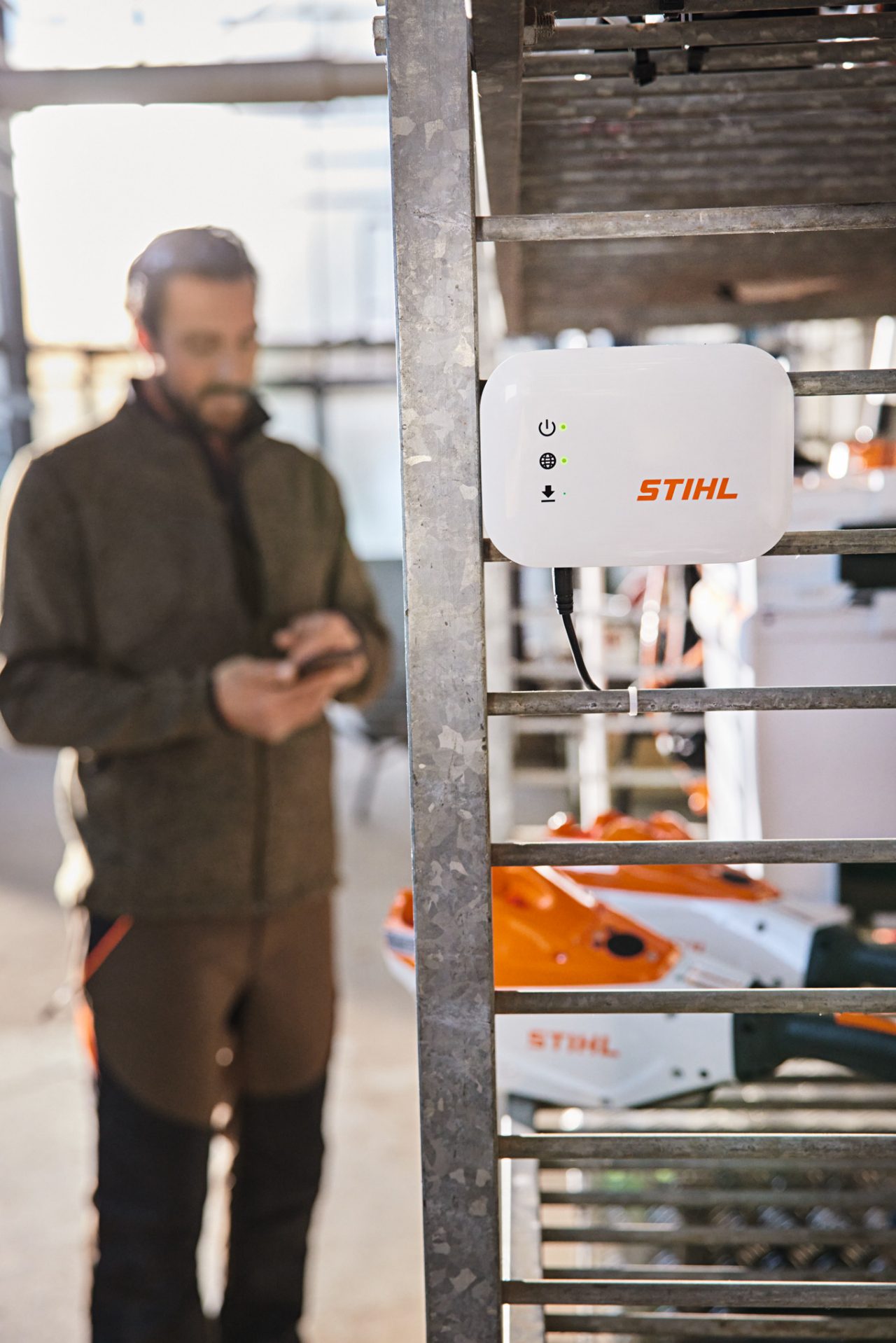 Une STIHL&nbsp;connected&nbsp;Box est suspendue à des étagères dans un atelier. En arrière-plan, un paysagiste interagit avec son smartphone. 