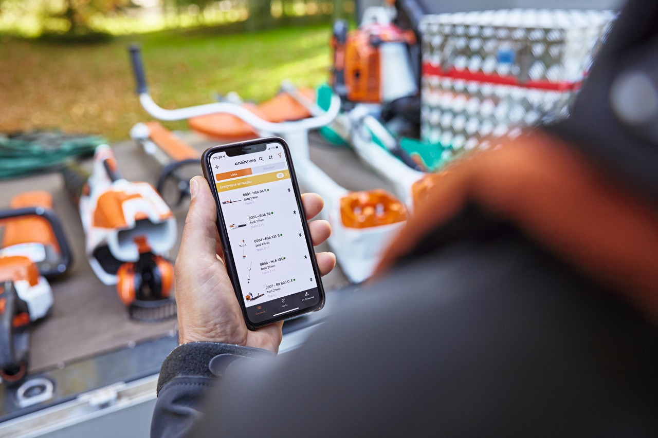 Une personne tient un smartphone dans sa main et consulte la liste des appareils dans l'application STIHL connected. Des appareils STIHL sont visibles en arrière-plan.