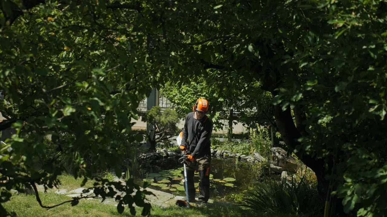 Achille lavora con un decespugliatore in giardino e indossa dispositivi di protezione STIHL, con un giardino e un laghetto circondato da vegetazione verde sullo sfondo, campagna Real People. STIHL People.